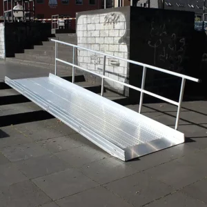 Eine barrierefreie Rampe aus Metall mit rutschfester Oberfläche, die zu einem erhöhten Gehweg führt. Die Rampe ist an einem städtischen Ort platziert, um Zugang für Rollstuhlfahrer zu ermöglichen.